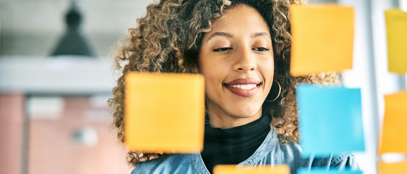 Auf dem Bild sieht man eine junge Frau, die lächelt und vor einer Glaswand, auf der Post-Its in gelb und blau sind, steht.