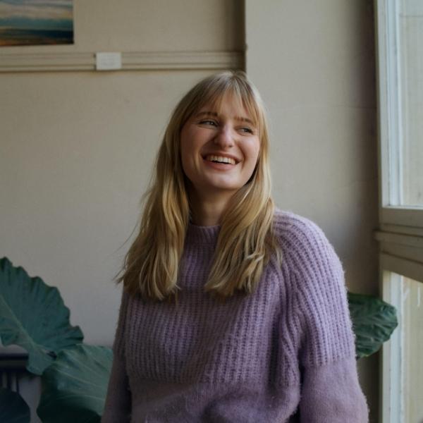 Charlotte von der Impact-Werkstatt steht in einem Treppenhaus vor einer großen Pflanze und lacht.
