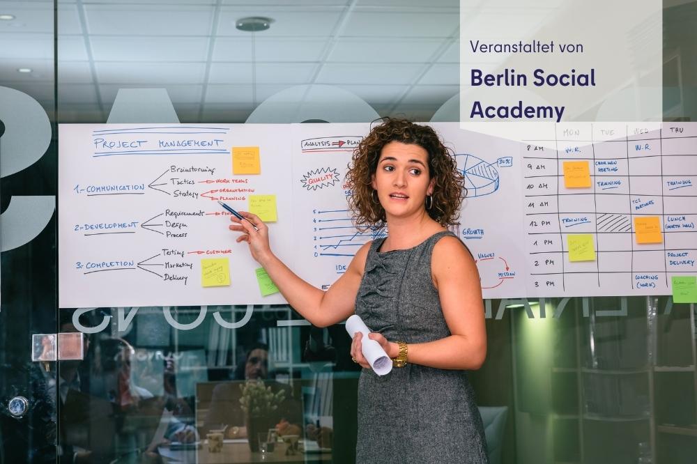 Auf diesem Bild sieht man eine Frau vor einem beschriebenen White-Board stehen, während sie etwas erklärt. Rechts oben steht geschrieben " Veranstaltet von: Berlin Social Academy"