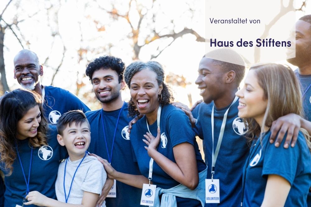 Auf diesem Bild sieht man eine Grupe fröhlicher Engagierter mit blauen T-Shirts, die sich umarmen. Rechts oben steht in blau geschrieben " veranstaltet von Haus des Stiftens".