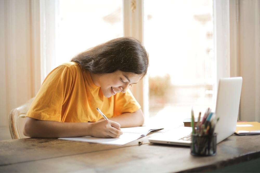 Auf diesem Bild sieht man eine junge Frau über ein Heft gebeugt etwas aufschreiben. Sie sitzt an einem Schreibtisch und vor ihr steht ein aufgeklappter Laptop.
