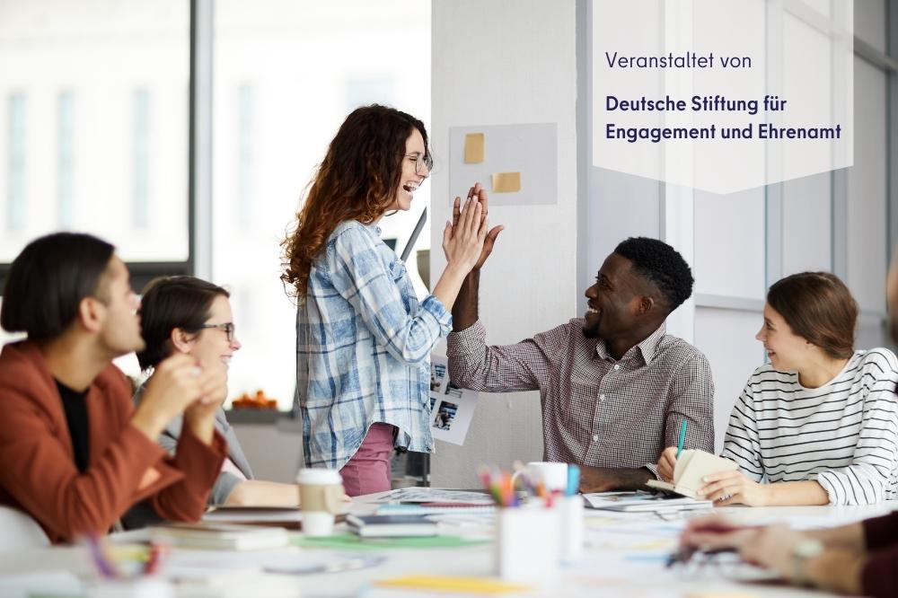 Auf diesem Bild sieht man eine Gruppe junger Leute in einem Seminarraum an einem großen Tisch. Im Hintergrund klatschen sich zwei ab. Oben rechts steht "Veranstaltet von Deutsche Stiftung für Engagement & Eherenamt" geschrieben.